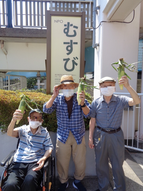 むすびさんの看板前でトウモロコシを掲げる